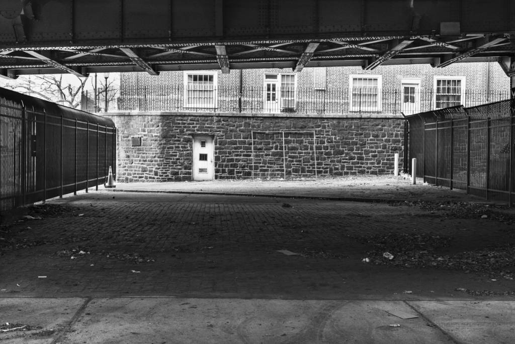 Black and white view of empty space under bridge, surrounded by steel fences and stone walls.