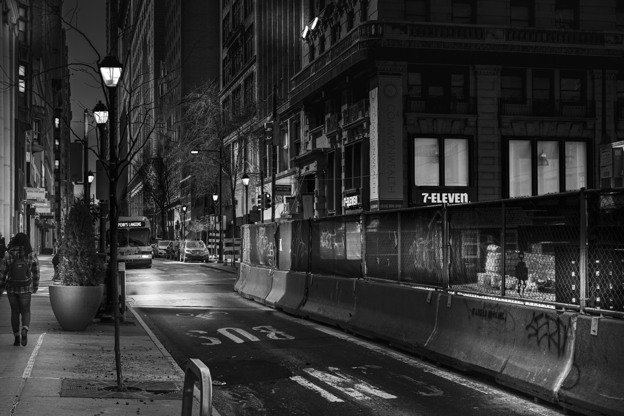 Black and white view at night of downtown commercial street with pedestrian and bus approaching.